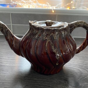 May include: A brown ceramic teapot with a lid. The teapot has a glossy finish with vertical, textured patterns. The handle and spout are also brown and textured. The teapot is designed for brewing tea.