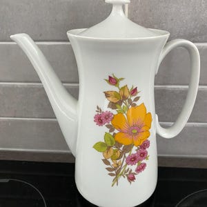 May include: White ceramic coffee pot with a lid and a curved handle. The pot is decorated with a floral design featuring a large yellow flower, pink flowers, and brown and green leaves. The pot has a long spout.