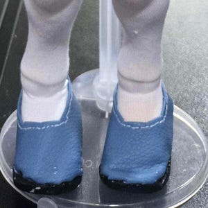 May include: A pair of blue doll shoes with a textured surface and visible light blue stitching along the top edge. The shoes are worn over white fabric socks and are positioned on a clear plastic circular doll stand base.