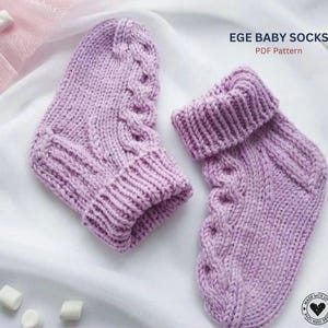 Könnte beinhalten: Zwei lavendelfarbene Babysocken, handgestrickt mit einem Zopfmuster auf der Vorderseite. Die Socken haben gerippte Bündchen und werden auf einer weißen Oberfläche präsentiert. Der Text auf dem Bild lautet "EGE BABY SOCKS PDF Pattern".