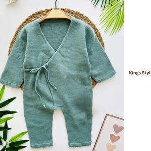 Puede incluir: Un pelele de bebé de punto verde salvia con mangas largas y un frente cruzado, atado con un cinturón de tela a juego. La prenda se exhibe en una percha de madera sobre un fondo tejido. El texto "Kings Style Romper" es visible.