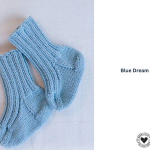 Könnte beinhalten: Ein Paar hellblaue Stricksocken mit gerippten Bündchen und einem strukturierten Design. Die Socken werden auf einer weißen Oberfläche präsentiert. Auf der rechten Seite steht "Blue Dream Socks".