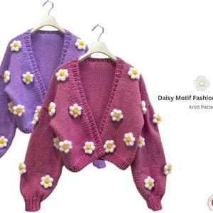 Könnte beinhalten: Zwei Daisy-Motiv-Mode-Cardigans, einer lila und einer rosa, hängen an Holzbügeln. Jeder Cardigan ist mit weißen und gelben Gänseblümchen-Applikationen verziert. Der Text "Daisy Motif Fashion Cardigan Knitt Pattern" ist sichtbar.