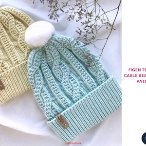 May include: Two cable-knit beanies with white pom-poms. One is light yellow, the other is light blue. Each has a ribbed brim and a small leather tag. The text "FIGEN TEXTURED CABLE BEANIE - KNIT PATTERN" is visible.