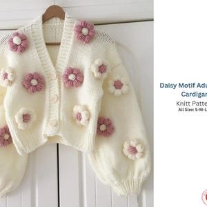 Könnte beinhalten: Cremefarbene, gecroppte Strickjacke mit Gänseblümchen-Verzierungen in Rosa und Weiß. Die Strickware hat einen V-Ausschnitt, einen Knopfverschluss und lange, puffige Ärmel. Der Text lautet "Daisy Motif Adult Crop Cardigan Knitt Pattern All Size: S-M-L-XL."