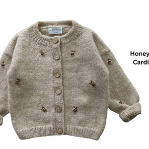 May include: Beige knit cardigan with embroidered honey bee details. Features a round neckline, button-up front, and ribbed cuffs. The text "Honey Bee Cardigan" is displayed on the right side of the image.