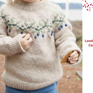 Pode incluir: Um suéter bege de malha com gola redonda e mangas compridas. O suéter apresenta um design verde e azul no peito e o texto "Lovely Winter Cardigan". O suéter é usado por uma criança.