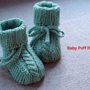 May include: A pair of knitted baby booties in a soft teal color. The booties have a cable knit design on the foot and a ribbed cuff. They are tied with matching teal yarn. The text "Baby Puff Romper" is visible in the image.