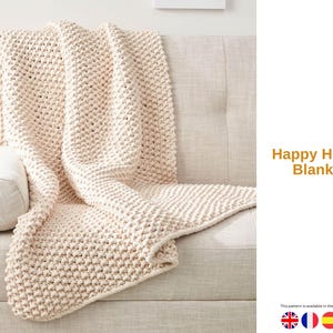 May include: A cream-colored, chunky knit blanket draped over a light-colored sofa. The blanket has a textured, raised pattern. The words "Happy House Blanket" are displayed in orange text. The blanket is a cozy home decor item.