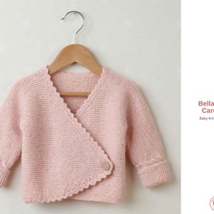 Pode incluir: Um cardigan rosa claro para bebé com um design cruzado, bordas onduladas e um fecho de botão único. As mangas têm punhos canelados. O texto "Bella Baby Cardigan" é visível no lado direito da imagem.