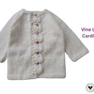 May include: Cream-colored knitted cardigan with long sleeves. The front features a row of decorative vine leaf appliques with purple button closures. The text "Vine Leaf Cardigan" is visible in the image.