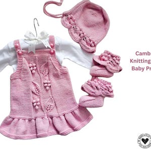 Peut inclure: Ensemble de bébé tricoté rose, comprenant une robe avec des détails de feuilles et de baies, un bonnet et des chaussons. La robe est superposée à un t-shirt blanc à manches longues. L'ensemble est présenté sur fond blanc. Texte : "Cambridge Knitting Set for Baby Princess."