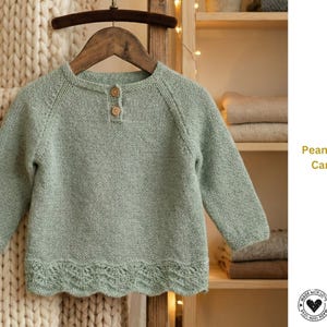 Puede incluir: Un cárdigan de bebé verde claro con mangas largas, dos botones de madera y un dobladillo festoneado. El suéter está colgado de una percha de madera. El texto "Peanet Baby Cardigan" es visible en el lado derecho de la imagen.