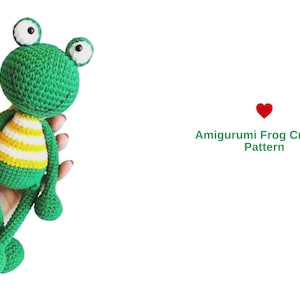 May include: A handmade green crochet frog amigurumi with large, round eyes and a striped yellow and white belly. The frog is a whimsical design, perfect for a child's toy or nursery decor. The image includes the text "Amigurumi Frog Crochet Pattern".