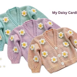 Peut inclure: Trois cardigans tricotés de couleurs pastel : bleu clair, lavande et pêche. Chaque cardigan est orné d'appliqués de marguerites blanches et jaunes. Le texte "My Daisy Cardigan" est visible. Les cardigans ont un col en V et une fermeture boutonnée.