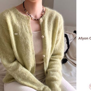 Puede incluir: Cárdigan verde claro con botones y textura suave y difusa. El cárdigan tiene cuello redondo y mangas largas. El texto "Afyon Cardigan" es visible en el lado derecho de la imagen. Se combina con una camiseta de color neutro.