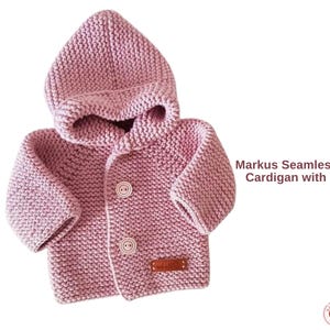 Könnte beinhalten: Hellrosa gestrickte Baby-Strickjacke mit Kapuze. Die Strickjacke hat zwei Knöpfe und ein kleines braunes Etikett. Der Text "Markus Seamless Baby Cardigan with Hood" ist auf der rechten Seite sichtbar.
