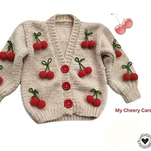 Peut inclure: Cardigan crème orné de cerises rouges et de tiges vertes. Le pull a un col en V, des boutons rouges et des poignets côtelés. Le texte "My Cheery Cardigan" est visible.