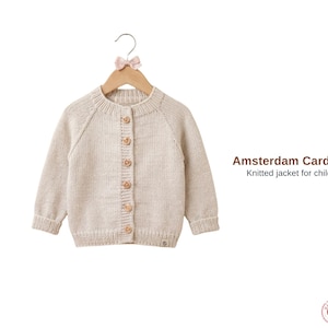 Puede incluir: Un cárdigan beige tejido para niños, colgado de una percha de madera con un lazo rosa. El cárdigan tiene cuello redondo, mangas largas y botones de madera. El texto dice "Amsterdam Cardigan, Chaqueta de punto para niños."