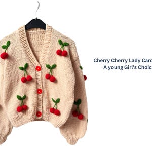 May include: A cream-colored, knitted cardigan adorned with red cherry appliqués and green leaves. The V-neck sweater has red buttons and is hanging on a black hanger. Text on the right reads "Cherry Cherry Lady Cardigan A young Girl's Choice."