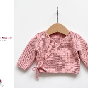 May include: A pale pink, knitted baby cardigan with long sleeves, displayed on a wooden hanger. The wrap-style sweater has a tie closure on the front. The text "Vargas Baby Cardigan Knitted Pattern" is on the left.