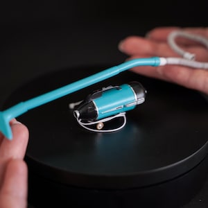 Può includere: Un mini aspirapolvere turchese e nero con un lungo manico turchese e un cavo bianco. L'aspirapolvere è appoggiato su una superficie nera. L'immagine è un primo piano.