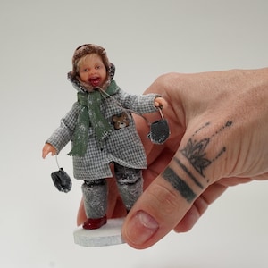 May include: A miniature doll of a child in winter attire. The doll is wearing a grey and white checked coat, a green scarf, a brown hat, and grey boots. The doll holds black mittens and has a small teddy bear in the coat pocket.