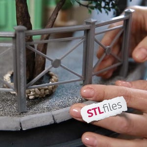 Puede incluir: Una valla gris en miniatura con un diseño en forma de X, parte de una escena de maqueta. La valla está sujeta por las manos de una persona. La escena incluye un árbol y una jardinera bordeada de piedras. El texto "STLfiles" es visible.