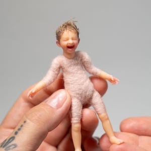 Könnte beinhalten: Eine Miniaturfigur eines lachenden Babys mit hellbraunem Haar, geschlossenen Augen und offenem Mund. Die Figur hat eine glatte, helle Hautoberfläche an Kopf und Gliedmaßen, während der Rumpf und die Arme mit einer weichen, flauschigen, blassrosa Textur bedeckt sind.