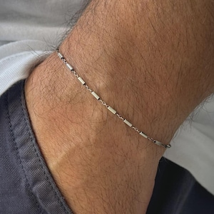 May include: A silver bracelet with a fine chain and small, rectangular bar links. The bracelet is worn on the wrist, highlighting its minimalist design. The jewellery is a simple accessory.