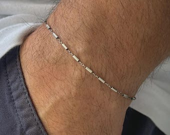 May include: A silver bracelet with a fine chain and small, rectangular bar links. The bracelet is worn on the wrist, highlighting its minimalist design. The jewellery is a simple accessory.