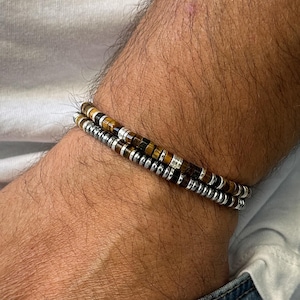 Może przedstawiać: Dwie bransoletki ułożone na nadgarstku. Górna bransoletka wykonana jest z brązowych i czarnych koralików z tygrysiego oka. Dolna bransoletka wykonana jest z srebrnych koralików.