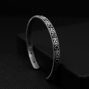 May include: A silver bangle bracelet with an intricate design featuring black accents. The bracelet is open and has a simple, elegant style.