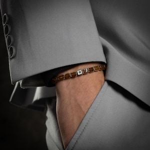 以下が含まれることがあります： 銀色のチャームが付いた茶色のビーズブレスレット。ブレスレットは手首に着用されています。