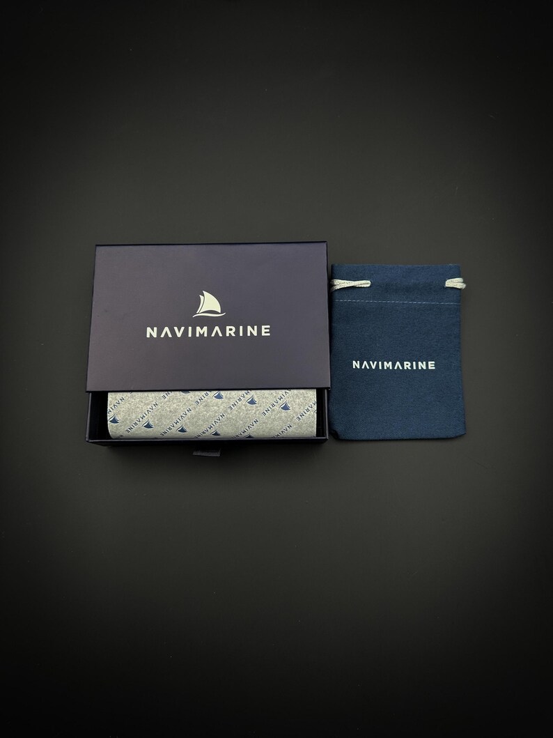 May include: A black box with the word "NAVIMARINE" printed on the top. The box is open and shows a patterned lining with the same logo. A small blue drawstring bag with the same logo is next to the box.