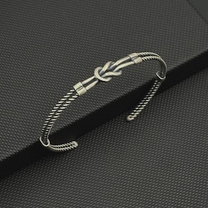Puede incluir: Una pulsera tipo brazalete de tono plateado con un diseño de nudo central. La banda está construida con dos hilos de alambre trenzado que se unen en el nudo, con detalles metálicos lisos que aseguran el punto de unión a ambos lados del nudo.