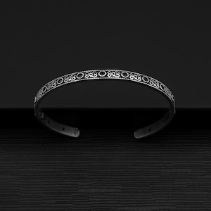 May include: A silver bangle bracelet with a decorative, intricate design. The bracelet is open and has a smooth, polished finish.
