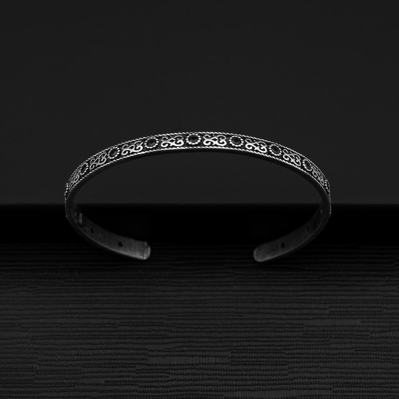 May include: A silver bangle bracelet with a decorative, intricate design. The bracelet is open and has a smooth, polished finish.
