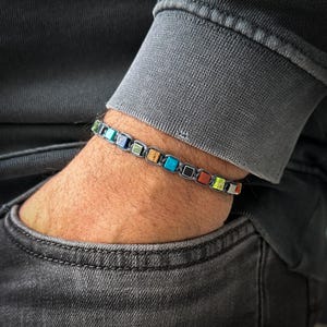 May include: A colorful bracelet with square beads in shades of blue, green, yellow, orange, and red. The bracelet is made with a gray cord and black accents.