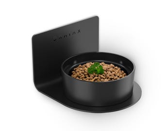 May include: A black elevated pet bowl with a round food container and a back support. The bowl is filled with dry pet food and garnished with a sprig of parsley. The word "DRAIAX" is printed on the back support.