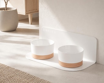May include: A white pet feeding station featuring two bowls with light wood bases, set on a white platform with a vertical back panel. The word DRALAX is embossed on the back panel. The bowls are white with a natural wood-toned bottom trim.