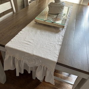 May include: A beige linen table runner with a ruffled edge and a circular embroidered design. A decorative serving tray with a glass top and a ceramic dish with a chicken lid sit on the runner, placed on a wooden table.