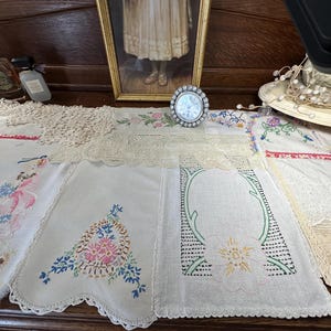 May include: A collection of white and off-white fabric linens featuring various embroidered floral patterns, intricate lace borders, and scalloped edges. The assortment includes rectangular pieces with colorful thread work, delicate crochet details, and openwork designs.