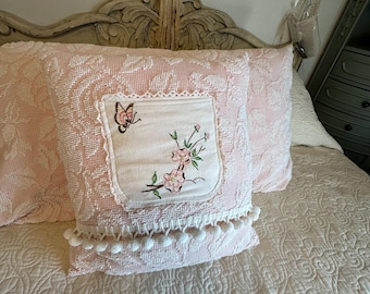 May include: A square throw pillow featuring a textured light pink fabric with a white lace-trimmed center patch. The patch displays an embroidered design of a small butterfly and pink flowers with green stems. A row of white pom-pom trim accents the bottom.