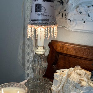 May include: A clear glass table lamp with a decorative base and a lampshade featuring a Paris-themed design. The lampshade has a pink trim and dangling beads. A lit candle sits nearby.