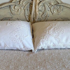 May include: Two white decorative pillows with floral patterns rest on a bed covered with a cream-colored, patterned quilt. The headboard is distressed white with carved details. The scene evokes a vintage, romantic bedroom aesthetic.