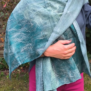 Könnte beinhalten: Ein türkis-blauer Schal mit einem strukturierten, aquarellartigen Muster. Der Schal ist über Schultern und Brust drapiert und erzeugt weiche Falten. Die Farbpalette umfasst Türkis-, Blau- und Weißtöne mit einem subtilen, fließenden Design. Der Schal scheint aus weichem, leichtem Material zu sein.