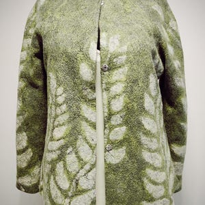 Könnte beinhalten: Grüne Filzjacke mit weißem Blättermuster. Die Jacke hat einen runden Kragen und wird mit drei silbernen Knöpfen geschlossen. Die Jacke wird auf einem schwarzen Mannequinkopf präsentiert und über einem weißen Kleidungsstück getragen.