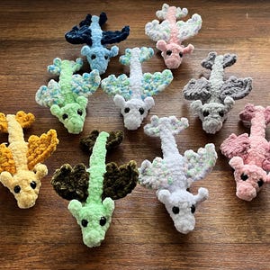 May include: A collection of ten crocheted dragon plushies in various colours, including blue, green, yellow, pink, and white. Each dragon has a unique design and features a long tail and wings.
