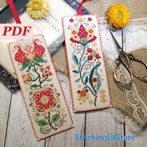 Op de afbeelding: Twee geborduurde bladwijzers met bloemmotieven. Eén heeft een grote rood-oranje bloem, de andere een blauw-rood bloemmotief. Beide hebben rood gestikte randen en een rood lint. Het woord "PDF" is zichtbaar.